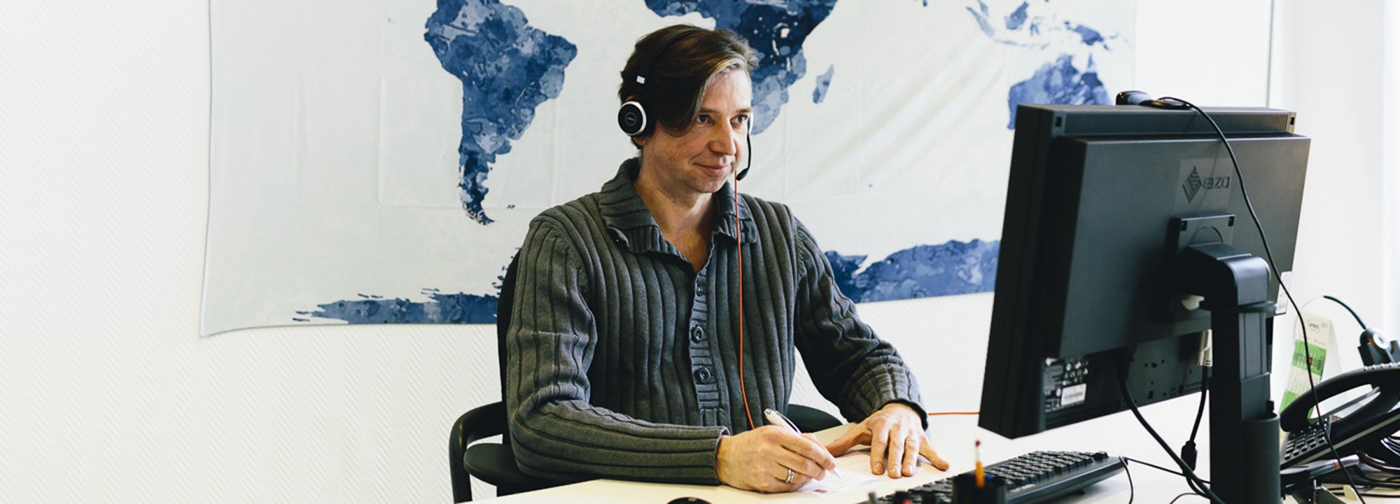 Mann mit Headset sitzt am Schreibtisch und blickt auf einen PC-Bildschirm.