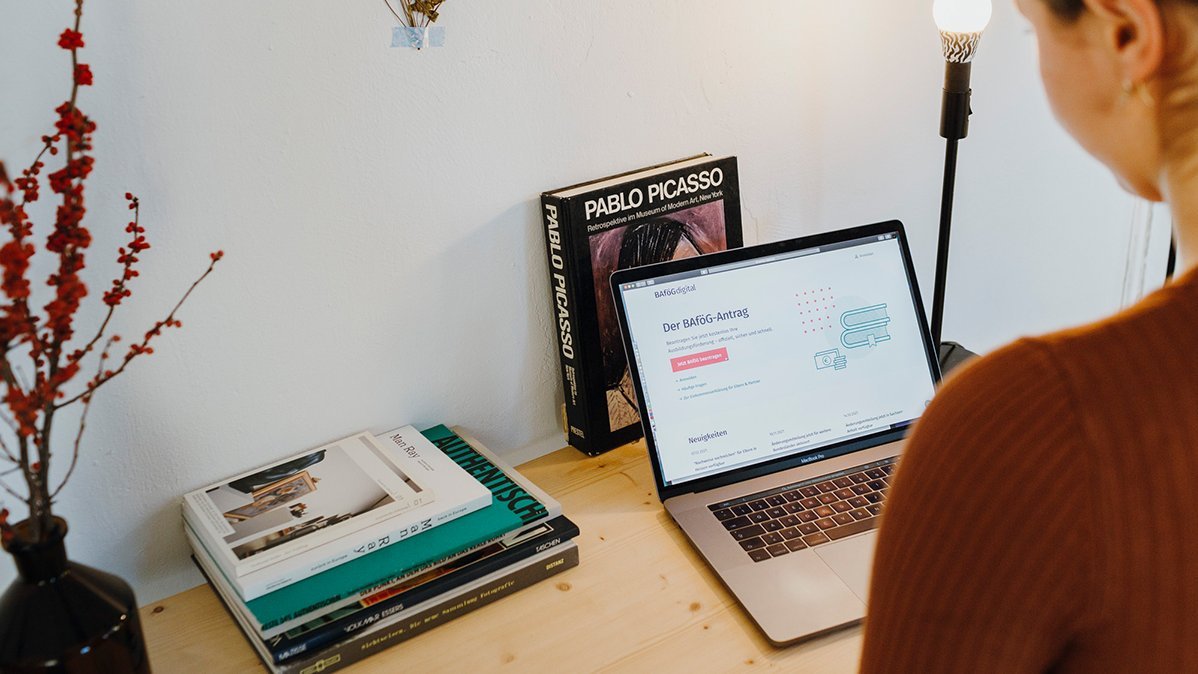 Frau, die auf den Bildschirm eines Laptops blickt. Auf dem Schreibtisch liegen Bücher.